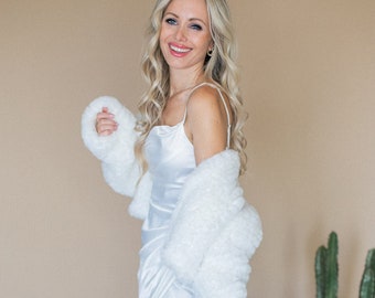 May include: A woman wearing a white satin slip dress with a white faux fur jacket. She is standing in front of a light brown background with potted plants.