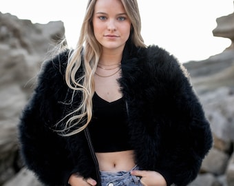 May include: A woman wearing a black faux fur bomber jacket with a zipper closure, a black crop top, and light blue jeans. She is standing in front of a rocky background.