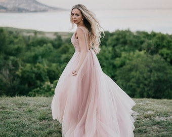 May include: A woman in a pink tulle gown with a beaded bodice stands in a field of green grass. She is looking over her shoulder and her hair is flowing in the wind.