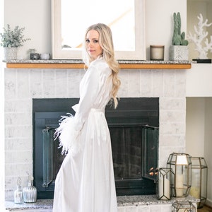 May include: A white satin robe with feather trim on the sleeves. The robe is worn by a person standing in front of a fireplace.