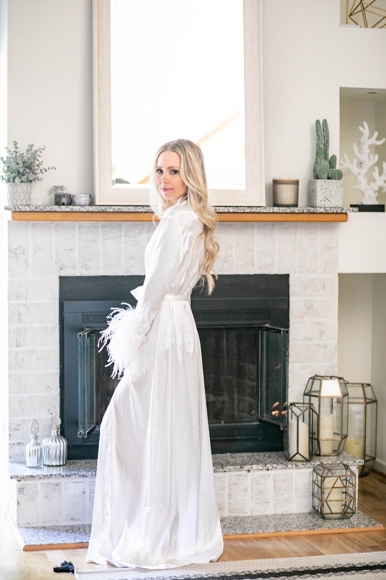 May include: A white satin robe with feather trim on the sleeves. The robe is worn by a person standing in front of a fireplace.