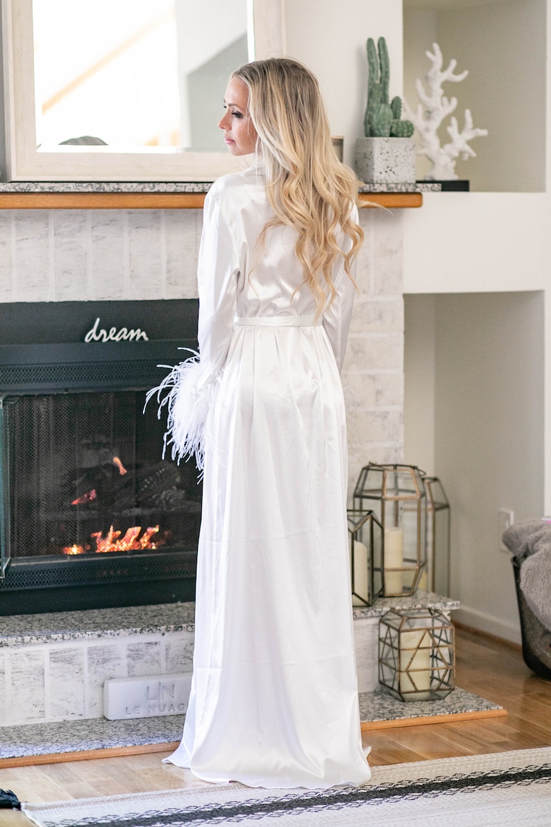 May include: A white satin robe with feather trim on the sleeves. The robe is tied at the waist and has a long, flowing skirt. The robe is worn by a woman with long blonde hair. The woman is standing in front of a fireplace with a sign that says "dream" above it.