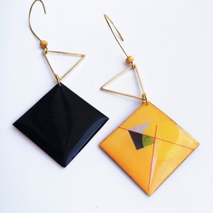 May include: A pair of geometric earrings featuring black and yellow resin squares with gold triangle accents. The earrings have a modern and abstract design.