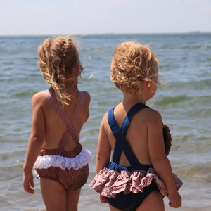 May include: Two young girls in ruffled swimsuits stand on a sandy beach, facing the ocean. The girl on the left is wearing a brown and white polka dot swimsuit with a white ruffle trim. The girl on the right is wearing a blue and white striped swimsuit with a pink ruffle trim.