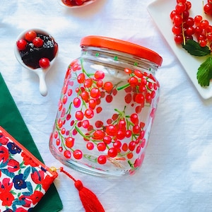 Puede incluir: Un tarro de vidrio transparente con tapa roja, pintado a mano con bayas rojas y tallos verdes. El tarro está lleno de una sustancia blanca.