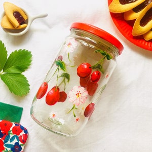 May include: A clear glass jar with a red lid, hand-painted with red cherries and white flowers. The jar is sitting on a white surface.