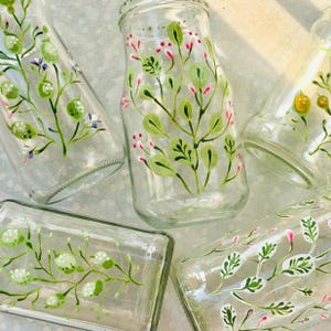 Può includere: Bottiglie e barattoli di vetro trasparente con disegni floreali dipinti a mano in verde, rosa e bianco. Le bottiglie hanno una varietà di forme, tra cui rotonde e rettangolari, e sono disposte in una ripresa ravvicinata.