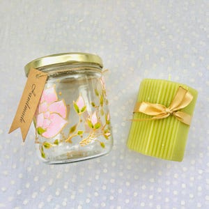 Peut inclure: Un bocal en verre avec un couvercle doré et des fleurs roses et des feuilles vertes peintes à la main. Le bocal est étiqueté "Handmade". Une bougie pilier verte avec un ruban doré est à côté du bocal.