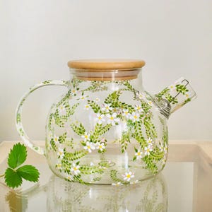 Puede incluir: Una tetera de vidrio transparente con un diseño floral. La tetera tiene una tapa de madera y un asa. El diseño floral presenta flores blancas y hojas verdes.