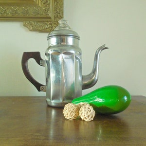 May include: A silver coffee pot with a brown handle and a green glass gourd on a wooden table. Two small, round, woven balls are in front of the gourd.