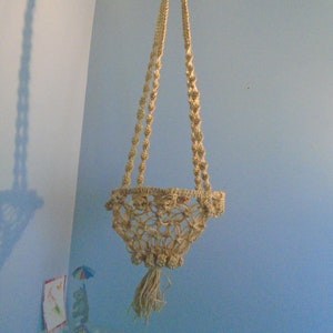 Peut inclure: Un porte-plante en macramé avec un design crocheté de couleur crème. Le porte-plante est suspendu à un crochet de plafond et a un pompon en bas.