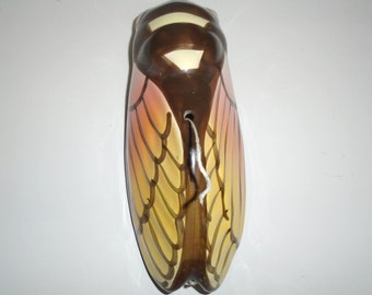 May include: A ceramic cicada figurine with brown, yellow, and pink painted details. The wings are detailed with a pattern of lines and the body has a small hole in the back.