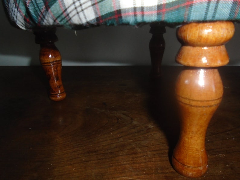 May include: Close-up of the wooden legs of a small stool with a plaid fabric top. The legs are turned and have a smooth, polished finish.