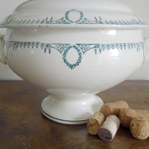 May include: A white ceramic tureen with a green and white floral design. The tureen has a lid and two handles. There are five cork bottle stoppers on the wooden surface next to the tureen.