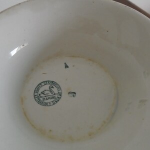 May include: Close-up of the bottom of a white ceramic bowl with a green circular stamp featuring a swan and the text "Usine de Ceramiques St. Amand - Tournai".