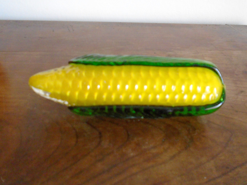 May include: A glass corn on the cob sculpture with a yellow cob and green husk.
