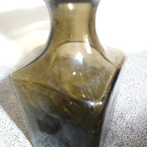 May include: A dark green glass bottle with a square base and a rounded neck. The bottle is empty and has a slightly cloudy appearance.