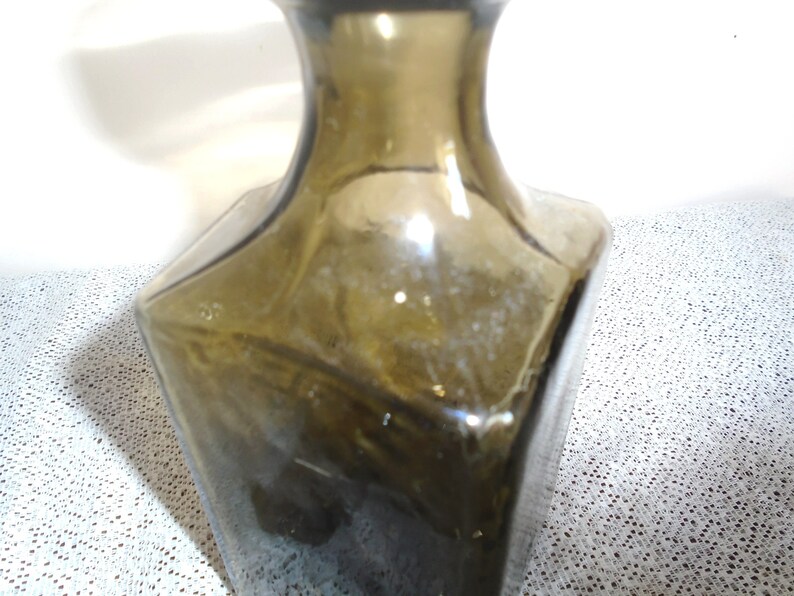 May include: A dark green glass bottle with a square base and a rounded neck. The bottle is empty and has a slightly cloudy appearance.