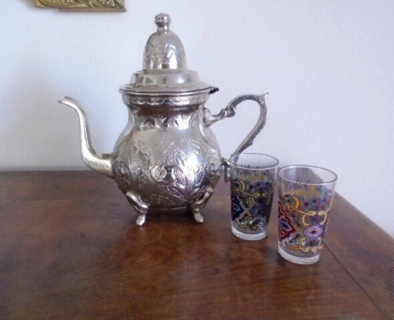 Puede incluir: Una tetera de plata con un dise&ntilde;o adornado y dos vasos con patrones coloridos. La tetera tiene una tapa y un pico. Los vasos est&aacute;n llenos de un l&iacute;quido transparente.