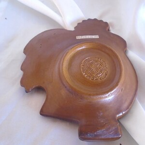 May include: A brown ceramic rooster-shaped dish with a circular indentation in the center. The bottom of the dish has a circular design with the text "Ave Maria Jesus" and a star in the center.