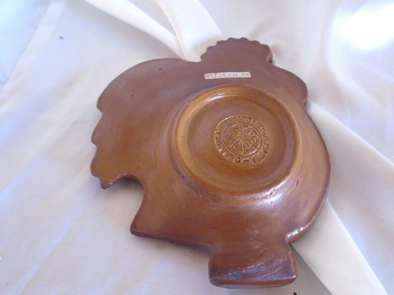 May include: A brown ceramic rooster-shaped dish with a circular indentation in the center. The bottom of the dish has a circular design with the text "Ave Maria Jesus" and a star in the center.