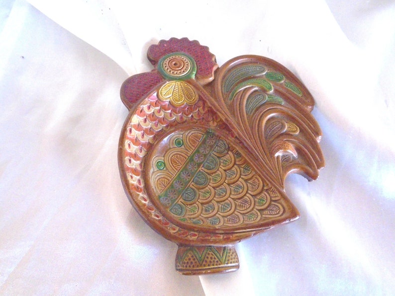 May include: A ceramic rooster figurine with a brown, red, green, and yellow painted design. The rooster has a detailed tail and a raised beak.