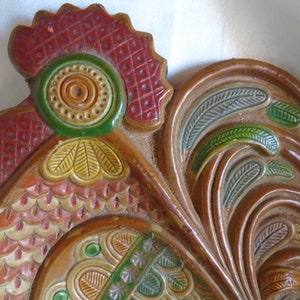 May include: A ceramic rooster wall decor with a red, green, and yellow painted design. The rooster has a detailed comb and wattle, and its feathers are painted in a stylized pattern.
