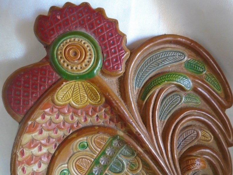 May include: A ceramic rooster wall decor with a red, green, and yellow painted design. The rooster has a detailed comb and wattle, and its feathers are painted in a stylized pattern.