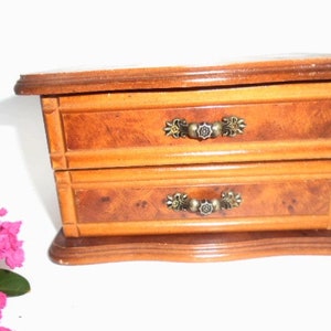 Peut inclure: Une bo&icirc;te &agrave; bijoux en bois avec deux tiroirs et des poign&eacute;es en m&eacute;tal orn&eacute;es. La bo&icirc;te est de couleur brun clair avec un motif de grain de bois.