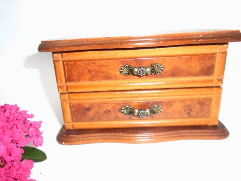 Peut inclure: Une bo&icirc;te &agrave; bijoux en bois avec deux tiroirs et des poign&eacute;es en m&eacute;tal orn&eacute;es. La bo&icirc;te est de couleur brun clair avec un motif de grain de bois.