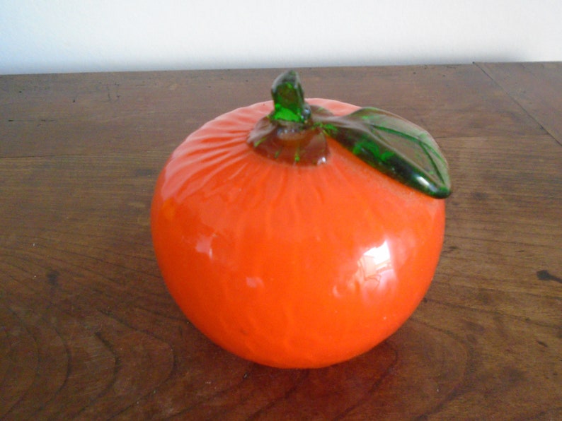 May include: A glass orange with a green glass leaf on top. The orange is a bright orange color and the leaf is a bright green color.