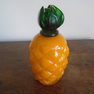 May include: A yellow glass pineapple-shaped bottle with a green glass top. The bottle has a textured surface that resembles a pineapple.