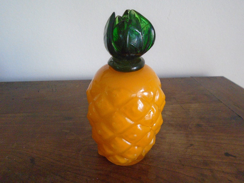 May include: A yellow glass pineapple-shaped bottle with a green glass top. The bottle has a textured surface that resembles a pineapple.