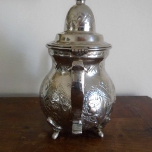 Puede incluir: Una tetera de plata con un dise&ntilde;o ornamentado y una tapa con bisagras. La tetera tiene tres patas y un asa. La tetera est&aacute; hecha de metal y tiene un acabado brillante.