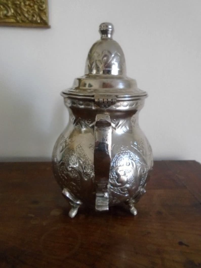 Puede incluir: Una tetera de plata con un dise&ntilde;o ornamentado y una tapa con bisagras. La tetera tiene tres patas y un asa. La tetera est&aacute; hecha de metal y tiene un acabado brillante.