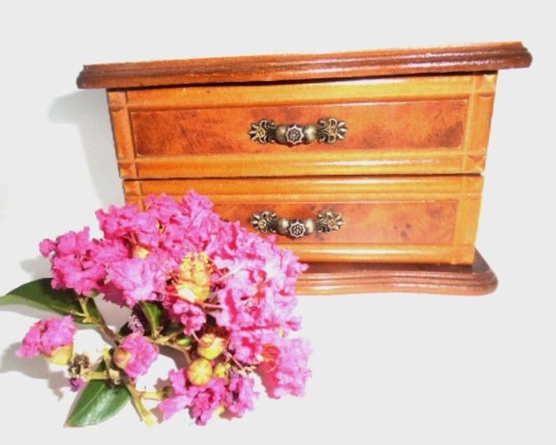 Peut inclure: Une bo&icirc;te &agrave; bijoux en bois avec deux tiroirs et des poign&eacute;es en m&eacute;tal orn&eacute;es. La bo&icirc;te est de couleur brun clair avec un motif de grain de bois. La bo&icirc;te est pos&eacute;e sur une surface blanche avec un bouquet de fleurs roses devant elle.