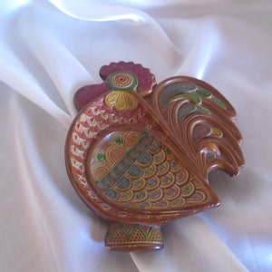 May include: A ceramic rooster dish with intricate details. The rooster is painted in shades of brown, orange, green, and yellow. The dish has a scalloped edge and a raised base.