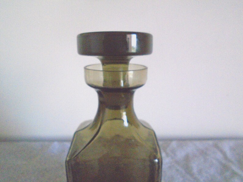 May include: A dark green glass bottle with a wide mouth and a removable stopper. The bottle has a square base and a rounded body.