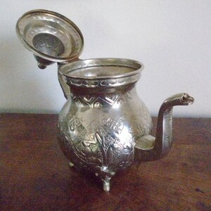 Puede incluir: Una tetera de plata con un dise&ntilde;o ornamentado y una tapa con bisagras. La tetera tiene un pico y un asa.