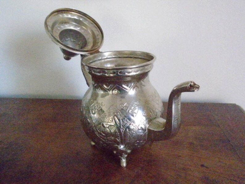 Puede incluir: Una tetera de plata con un dise&ntilde;o ornamentado y una tapa con bisagras. La tetera tiene un pico y un asa.