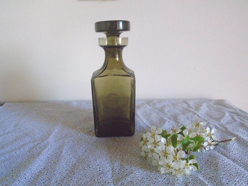 May include: A dark brown glass decanter with a stopper. The decanter is sitting on a white tablecloth with a sprig of white flowers.