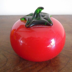 May include: A red glass tomato with a green stem and a black leaf. The tomato is sitting on a wooden surface.