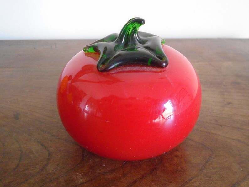 May include: A red glass tomato with a green stem and a black leaf. The tomato is sitting on a wooden surface.