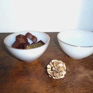 May include: Two white ceramic bowls with a gold rim. One bowl is filled with pieces of dark chocolate. The bowls are on a wooden surface.
