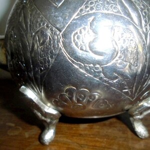 Puede incluir: Tetera de plata con intrincados dise&ntilde;os florales y de p&aacute;jaros, con tres patas y un pico. La tetera tiene un acabado brillante y est&aacute; decorada con un dise&ntilde;o en relieve.