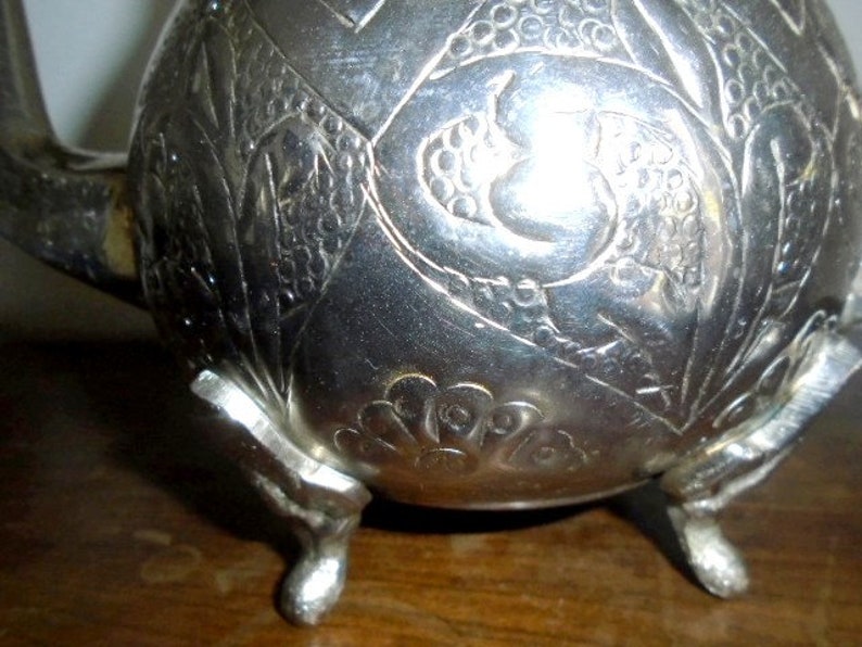 Puede incluir: Tetera de plata con intrincados dise&ntilde;os florales y de p&aacute;jaros, con tres patas y un pico. La tetera tiene un acabado brillante y est&aacute; decorada con un dise&ntilde;o en relieve.