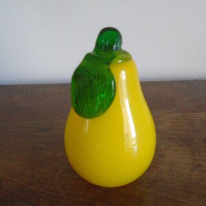 May include: A yellow glass pear with a green glass leaf on top. The pear is sitting on a wooden surface.