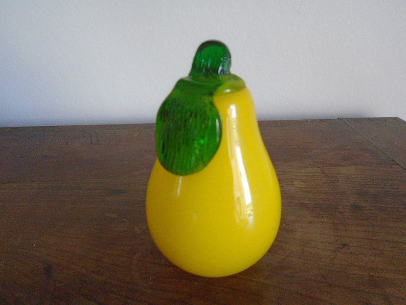 May include: A yellow glass pear with a green glass leaf on top. The pear is sitting on a wooden surface.
