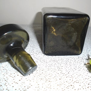 May include: A dark green glass bottle stopper with a round base and a cylindrical top. The stopper is separated from a square, dark green glass bottle.