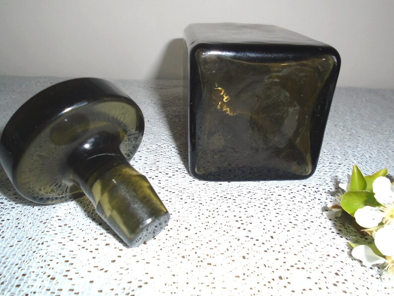 May include: A dark green glass bottle stopper with a round base and a cylindrical top. The stopper is separated from a square, dark green glass bottle.